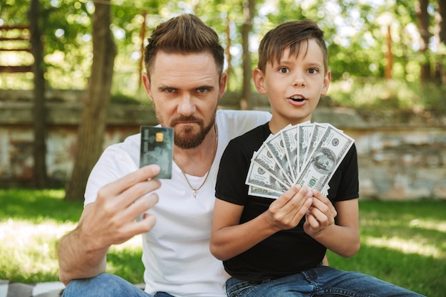 Padre joven serio sentado con su pequeño hijo con dinero y tarjeta de crédito.