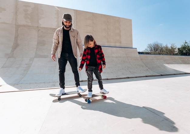 Foto padre joven y su hijo andan en patineta en un parque de patinaje con toboganes afuera en el día soleado.