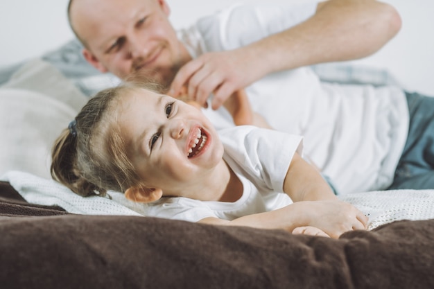 El padre juega con la pequeña hija 2-4 en la cama. Papá le hace cosquillas a los pies de los niños. Familia, divirtiéndose