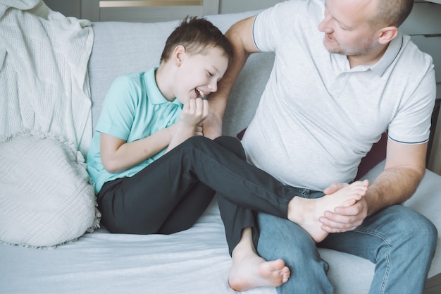 Padre juega con su hijo 7-10 sentado en el sofá. Papá le hace cosquillas a los pies de los niños. Familia, divirtiéndose