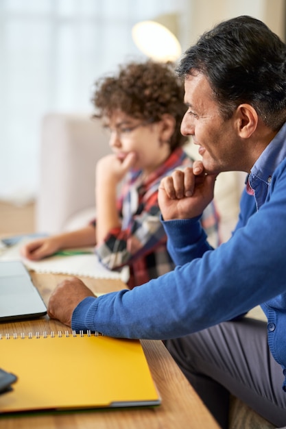 Foto padre latino de mediana edad revisando la tarea, ayudando a su lindo hijo, colegial con estudios mientras está sentado en la mesa en casa. estudio remoto, familia, concepto de paternidad