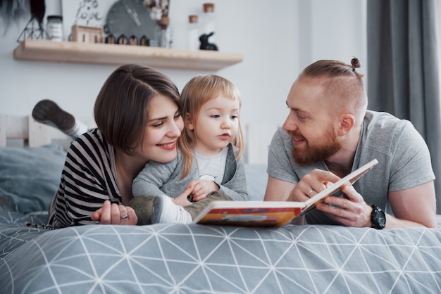 Padre, madre e hija leyendo el libro infantil en un sofá en la sala de estar. La gran familia feliz leyó un libro interesante en un día festivo. Los padres aman a sus hijos