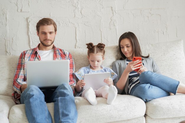 Padre, madre e hija usando dispositivos electrónicos sentados en el sofá en la sala de estar.