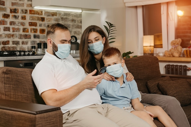 Padre, madre e hijo están sentados en un sofá con mascarillas médicas para evitar la propagación del coronavirus (COVID-19). Familia durante la cuarentena en casa. Los familiares están leyendo noticias en un teléfono inteligente en un sofá