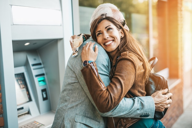 Padre mayor y su hija usando un cajero automático juntos para retirar dinero. Son felices. Día soleado.