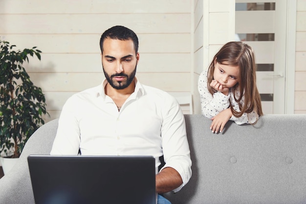 Padre multicultural barbudo que trabaja con una computadora portátil en la cómoda sala de estar, su pequeña hija observa cómo trabaja el padre