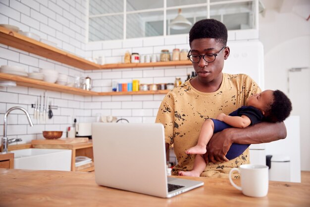 El padre multitarea tiene un hijo bebé dormido y trabaja en una computadora portátil en la cocina