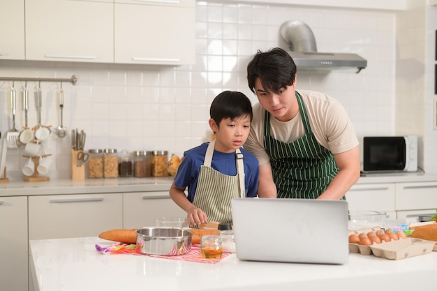Un padre y un niño asiáticos felices están usando una computadora portátil para aprender a cocinar