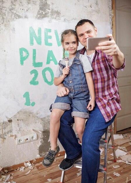 Padre con niños planeando año nuevo con lista de verificación en la pared y haciendo selfie con teléfono inteligente