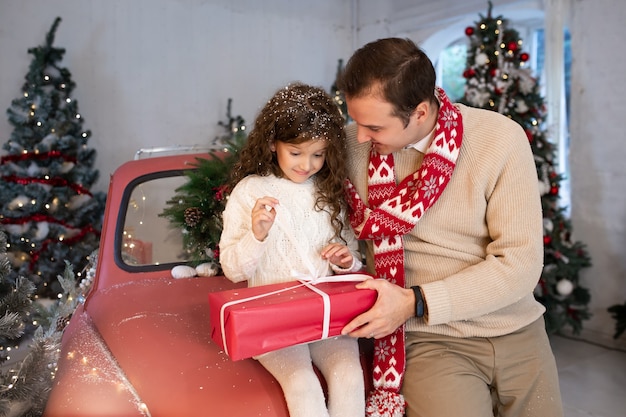 Padre presenta a su hija caja de regalo cerca del árbol de Navidad y el coche rojo.