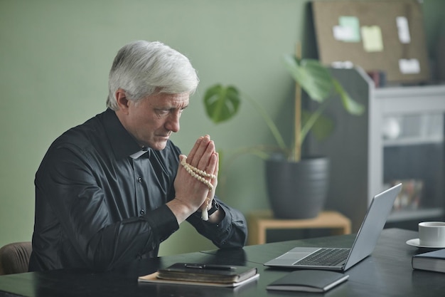 Padre sênior de cabelos brancos segurando rosário rezando na mesa no escritório