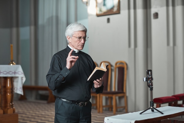 Foto padre sênior lendo a bíblia e filmando sua performance em um telefone celular enquanto está na igreja