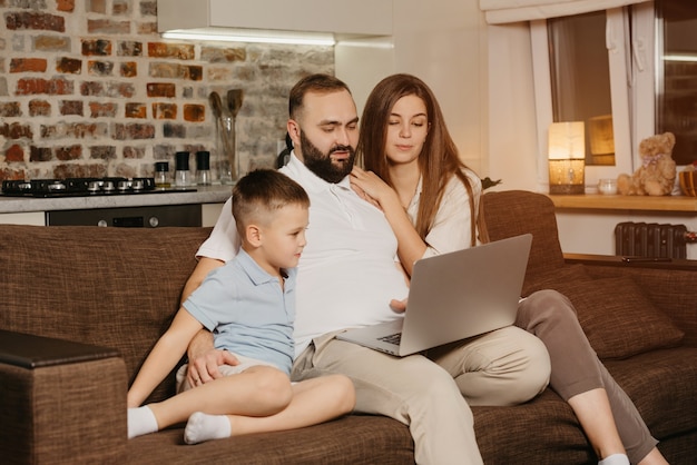 Un padre serio con barba está tratando de trabajar de forma remota en una computadora portátil cerca de su hijo y una esposa curiosa en casa