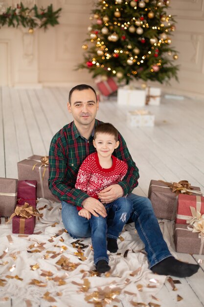 Padre sonriente abraza a su hijo en el árbol de Navidad, rodeado de regalos. Felices vacaciones en familia.