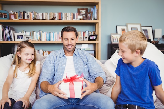 Padre sonriente con caja de regalo con niños sentados en el sofá