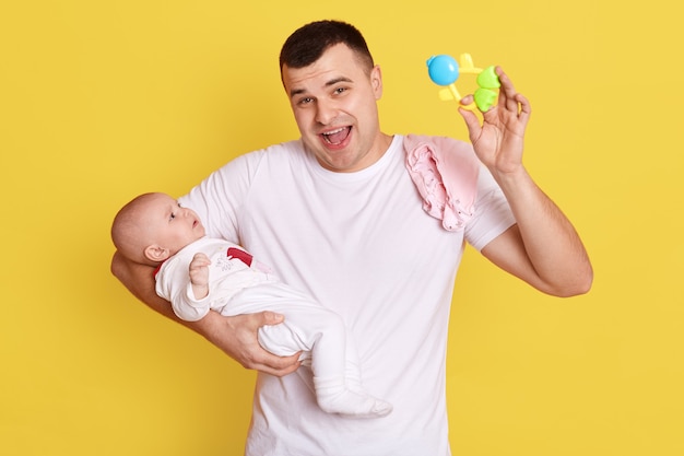 Padre sosteniendo un juguete puf y animando al bebé en su mano, posando aislado sobre una pared amarilla, feliz papá guapo gritando con camiseta blanca casual jugando con una niña.