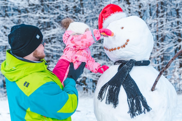 Padre sosteniendo a su hija en las manos ayudándola a poner nariz de zanahoria al concepto de muñeco de nieve