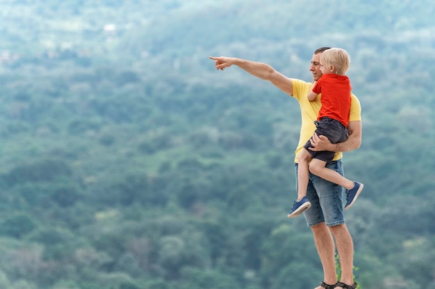 Padre sostiene a su hijo en sus brazos y señala con su dedo en algún lugar en la distancia Padre e hijo en una colina en las montañas