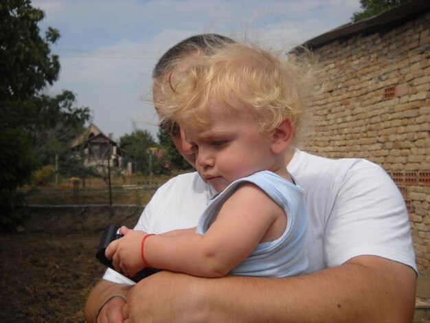 El padre sostiene en sus brazos a un niño pequeño con cabello rubio y ojos azules El niño mira hacia abajo con el ceño fruncido pensativo Verano en el pueblo