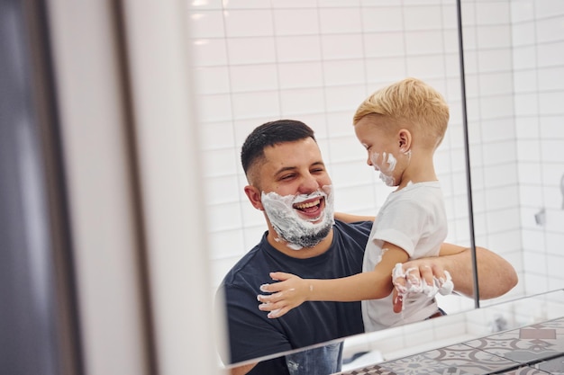 Padre con su hijo está en el baño diviértase usando gel de afeitar y mirándose en el espejo.