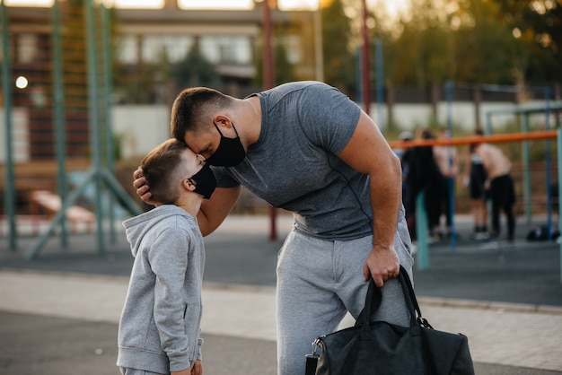 Un padre y su hijo se paran en un campo de deportes con máscaras después del entrenamiento durante la puesta de sol.