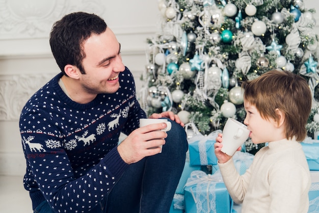 Padre con su hijo sentado cerca del árbol de Navidad y bebiendo té caliente