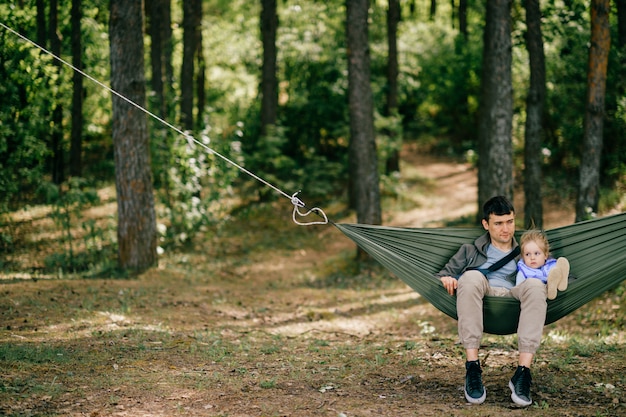 Padre con su pequeña niña artística divertida niña descansando y jugando en la naturaleza. Padres adultos de sexo masculino con la hija hermosa joven que miente en hamaca.