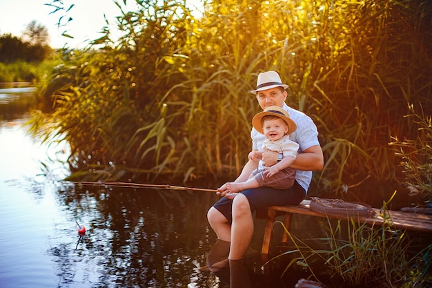 Padre con su pequeño hijo están pescando sentado en un muelle de madera junto al estanque.