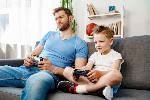 Padre y su pequeño hijo jugando videojuegos juntos en el sofá en casa