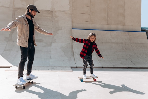 Padre y su pequeño hijo vestidos con ropa casual andan en patineta en un parque de patinaje con toboganes afuera en el día soleado.