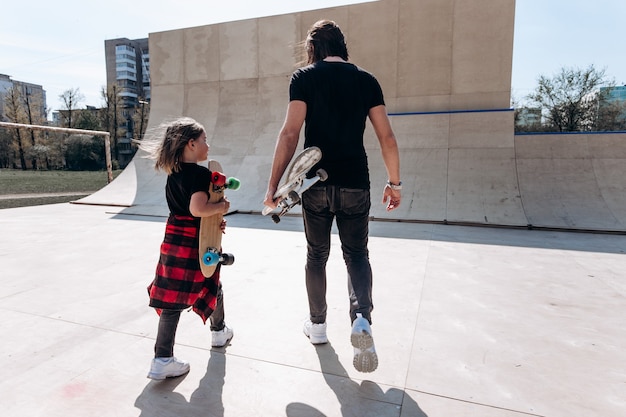 Padre y su pequeño hijo vestidos con ropa casual elegante caminan con las patinetas en sus manos en un parque de patinaje en el día soleado.