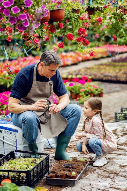 Padre trabajando en un centro de jardinería y enseñando a su pequeña hija a plantar flores en una maceta