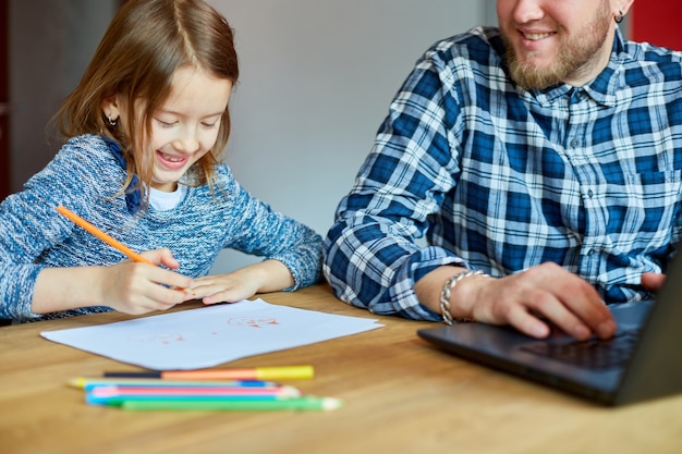 Padre trabajando en su oficina en casa en una computadora portátil, su hija se sienta junto a ella y dibuja