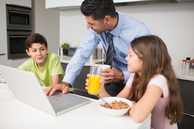 Padre usando laptop y niños desayunando en la cocina