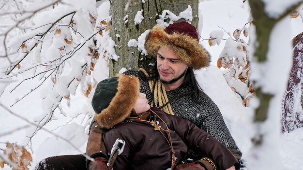 Padre vikingo y su hijo se sienta y habla sobre la nieve en el bosque de invierno