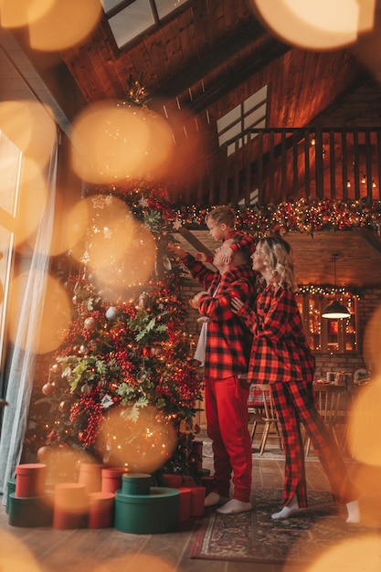 Foto los padres activos sonrientes con el hijo pequeño en trajes de pijama a cuadros rojos esperando a papá noel en el interior