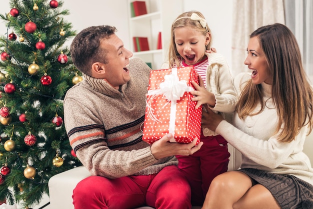 Los padres alegres sorprenden a su linda niñita con un regalo para Navidad o Año Nuevo.