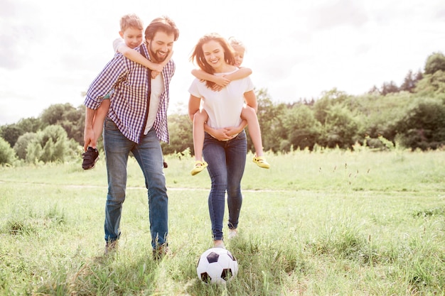 Los padres caminan por el prado y sostienen a los niños a la espalda. La mujer está jugando con la pelota en el césped con la pierna. hombre y mujer están mirando la pelota.
