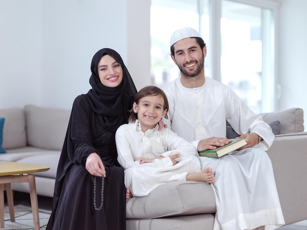 Padres de familia musulmanes tradicionales con niños leyendo Corán y rezando juntos en el sofá antes de la cena iftar durante una fiesta de ramadán en casa