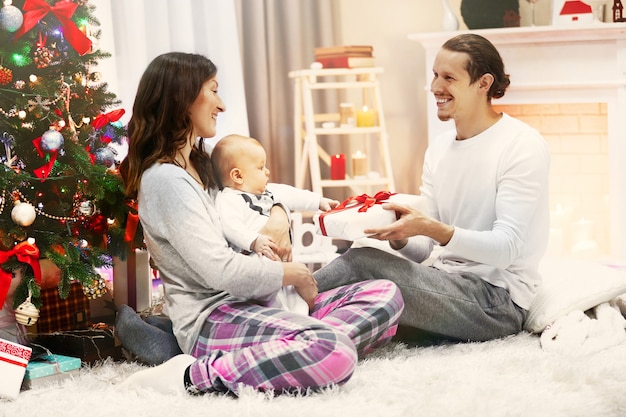 Padres felices con el bebé cerca del árbol de Navidad en el piso de la habitación decorada