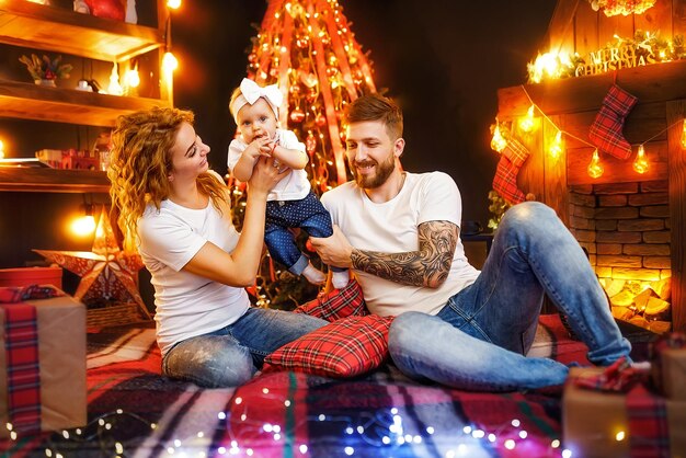 Padres felices jugando con su hija mientras se sientan juntos cerca del árbol de Navidad y la chimenea