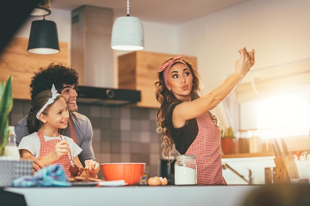 Foto los padres felices y su hija están preparando galletas juntos en la cocina. están tomando selfie con smartphone.