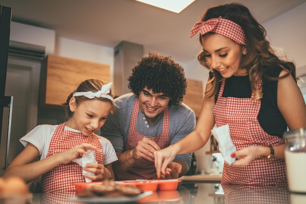 Los padres felices y su hija están preparando galletas juntos en la cocina. La niña ayuda a sus padres a poner migas de chocolate en las galletas.