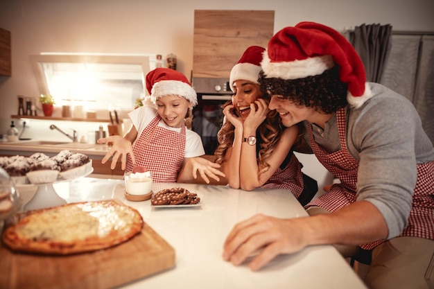 Los padres felices y su hija con gorra de Papá Noel están preparando la comida juntos en la cocina. La niña está ofreciendo el almuerzo a su madre y a su padre.