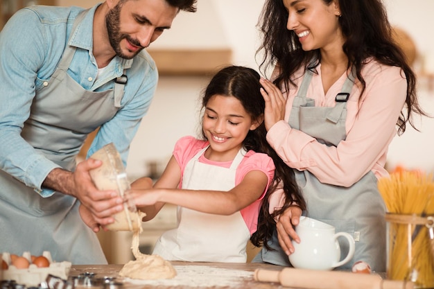 Padres felices y su pequeña hija amasando masa en la cocina, madre y padre enseñando a las mujeres