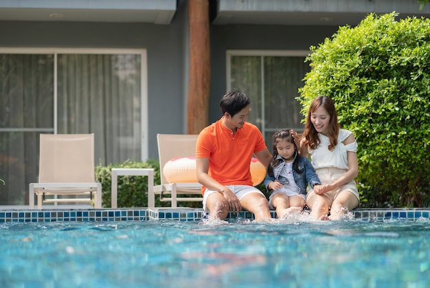 Los padres y la hija usan sus piernas para salpicar agua en el borde de la piscina mientras se relajan en el hotel en verano.