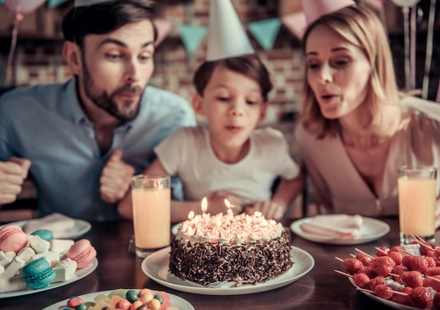 Los padres y el hijo felices están soplando velas en el pastel mientras están sentados a la mesa en la cocina decorada durante la celebración del cumpleaños