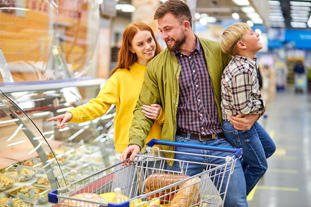 Padres con hijo inquieto en la tienda de comestibles, mujer eligiendo comida para la cena mientras su esposo sostiene a su hijo en las manos, cerca de vitrinas con comida