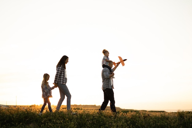 Los padres con hijos caminan en el verano al atardecer caminando por el campo Papá lleva a un bebé con un avión sobre sus hombros Mamá lleva a su hija de la mano Familia adoptiva