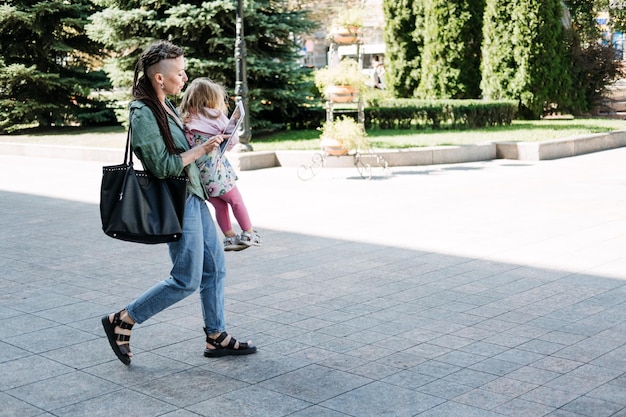 Padres independientes ocupados mamá trabajadora con una hija pequeña en las manos caminando y mirando la tableta en